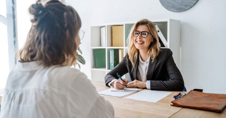 A smiling white woman with wavy blond hair and glasses sits across a table from a brown haired woman she is interviewing